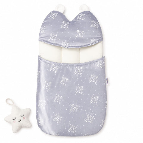 A baby footmuff in a light lavender-gray satin-like fabric with a white floral and star pattern, featuring a white inner lining and two white pom-poms. A small white plush star toy with embroidered facial features is placed to the left on a clean white ba