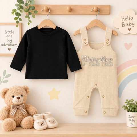 Baby clothing set displayed on wooden hangers against a nursery-style wall, featuring a black long-sleeve top and a cream ribbed overall romper with 'Paris' in rhinestones. A teddy bear, small booties, and decorative wall signs surround the outfit.