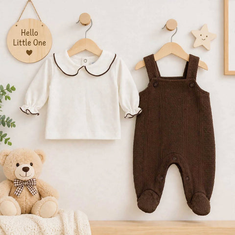 A baby outfit display on wooden hangers against a light wall, featuring a white long-sleeve top with a dark brown Peter Pan collar and cuffs, and a dark brown footed overall romper. A teddy bear sits on a blanket at the lower left, with decorative wall ha