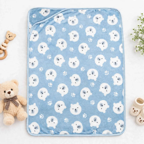 A light blue baby blanket with a repeating pattern of white polar bear faces and paw prints, styled in a flat-lay with a teddy bear, rattle, and baby shoes.