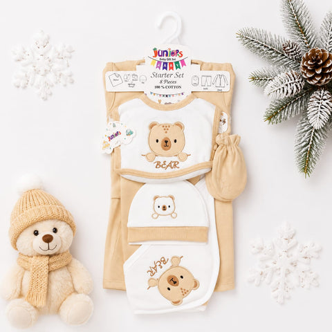 A baby starter set laid flat on a white background with holiday styling props. The set includes a cream and white bib with a teddy bear graphic, matching cream and white footmuff, cream and white booties, and a cream and white hat, all from Junior's Party