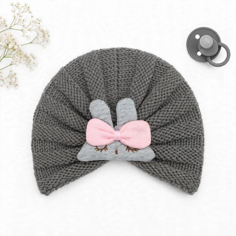 A gray knit baby hat with a bunny face applique and pink bow, shown on a white background with a pacifier and baby's breath flowers as props.