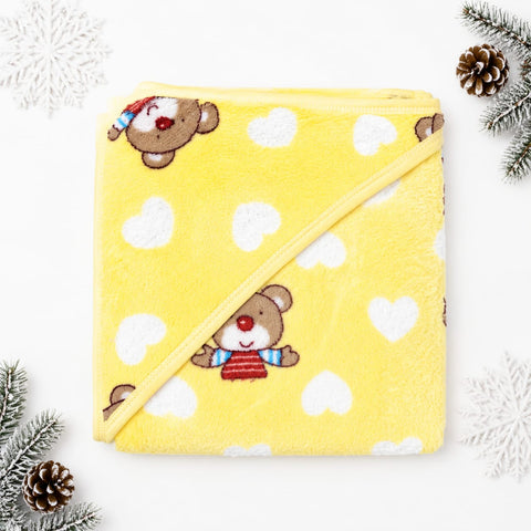 A yellow plush hooded towel blanket with a diagonal strap, decorated with white hearts and cartoon teddy bears wearing red and blue striped sweaters. The towel is styled in a flat-lay with pine branches and white snowflake decorations on a white backgroun