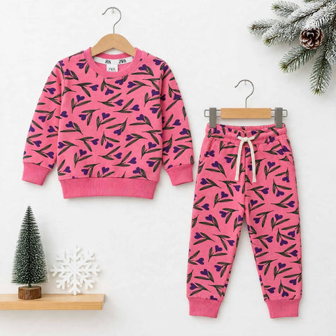 A children's two-piece outfit set consisting of a pink long-sleeve sweatshirt and matching jogger pants, both featuring a dark green leaf pattern with purple heart accents. The sweatshirt is shown on a wooden hanger on the left, and the pants on a metal h