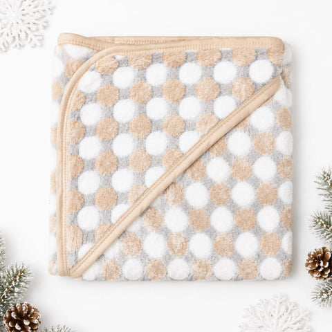 A folded plush hooded blanket with a beige trim, featuring a pattern of large beige and white polka dots on a light gray background, styled with holiday decorations on a white surface.