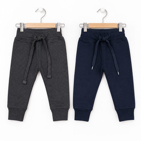 Two pairs of children's jogger sweatpants hanging on wooden hangers against a white background. The pair on the left is dark heather gray with a drawstring waist, and the pair on the right is dark navy blue with a drawstring waist.
