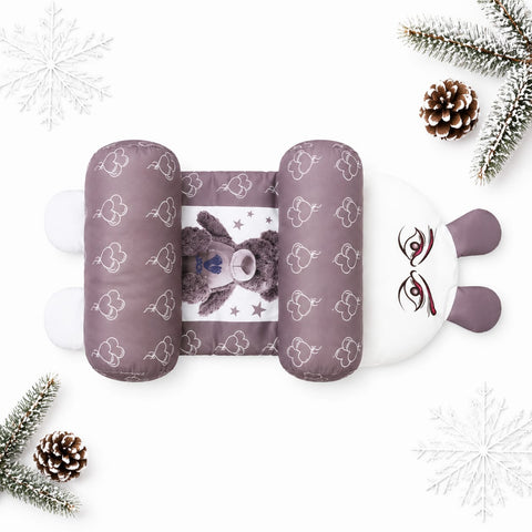 A plush baby support pillow styled in a flat lay with Christmas-themed props. The pillow features a dusty purple bunny face with white cloud patterns, a white inner support section, and a brown teddy bear plush in the center.
