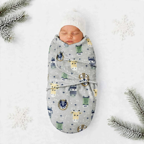 A sleeping newborn baby wrapped in a light gray blanket with a colorful animal print, wearing a white knit beanie, laid on a white background with decorative snowflakes and pine branches.