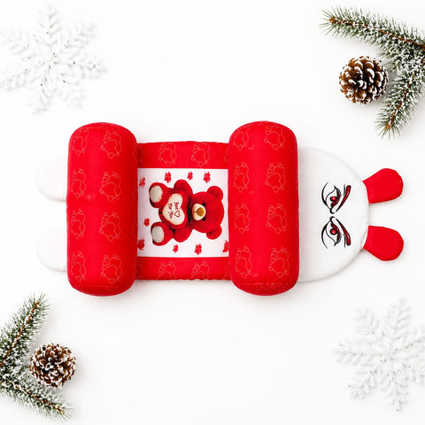 A red and white baby head and neck support pillow styled in a flat lay with white snowflakes, pinecones, and green pine branches on a white background.