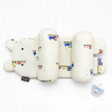 A baby support pillow designed in the shape of a bear, featuring a cream-colored fabric with a colorful cartoon train pattern. A small black fabric tag is attached near the bear's nose, and a blue pacifier is placed to the right on a clean white backgroun