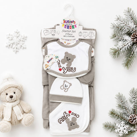 A baby starter set including a long-sleeve romper, bib, and hat, displayed on a hanger with decorative props like a teddy bear and pine branches on a white background.