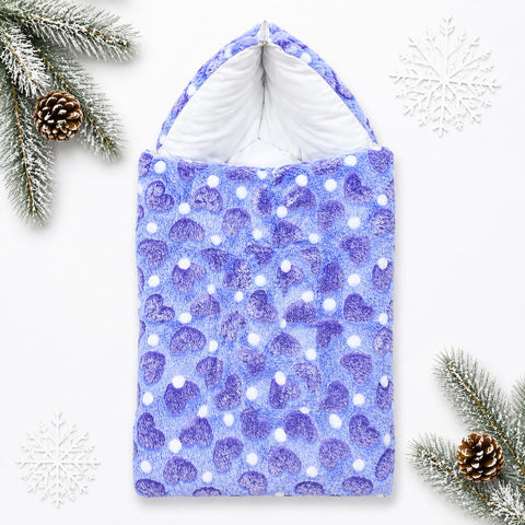 A plush hooded blanket or pouch in a light blue and purple watercolor pattern with white polka dots, styled flat-lay on a white background with pine branches, snowflakes, and pinecones.