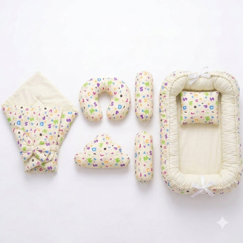 A baby bedding set laid out on a white background, including a quilted blanket with a colorful alphabet print and bow, a U-shaped pillow, two cylindrical pillows, a cloud-shaped pillow, and a bassinet cover with matching printed fabric and white ribbon ti