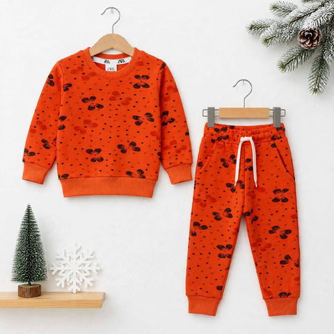 A children's two-piece outfit set consisting of an orange sweatshirt and matching jogger pants, both printed with small black motifs and dotted patterns. The sweatshirt is shown on a wooden hanger on the left, and the pants on a gray hanger on the right, 