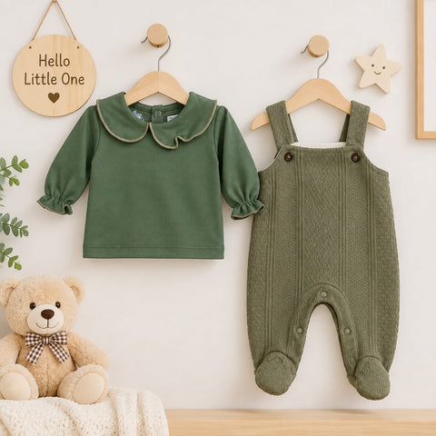 A baby clothing set displayed on wooden hangers against a light wall, featuring an olive green long-sleeve top with a ruffled Peter Pan collar and matching olive green footed overalls. A teddy bear and decorative wall hangings are visible in the styled sc