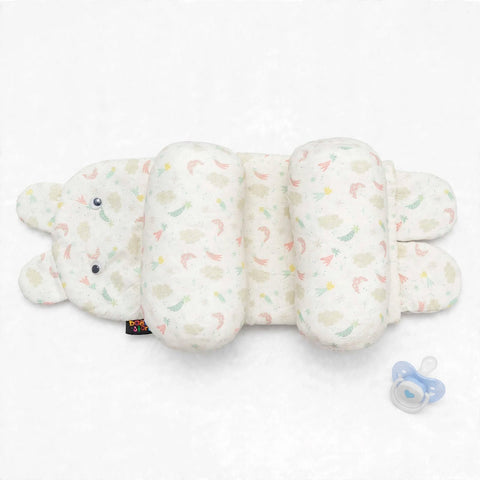 A plush baby support pillow shaped like a bunny, featuring a soft cream-colored fabric with a pastel rainbow pattern of rainbows and clouds. A small black fabric tag is visible near the bunny's nose, and a blue pacifier lies to the right on a clean white 