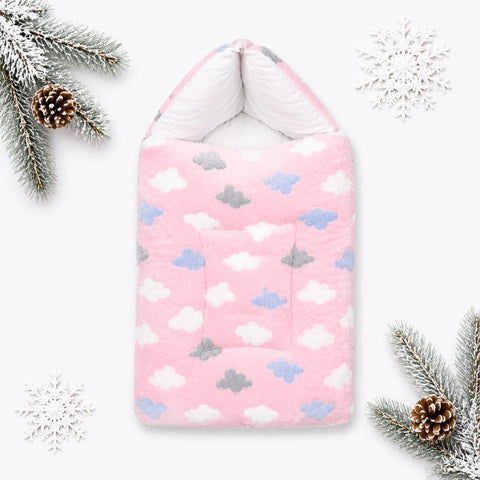 A pink quilted baby blanket or swaddle with a cloud pattern in white, light blue, and gray, styled with winter-themed props like pine branches, pinecones, and snowflakes on a white background.