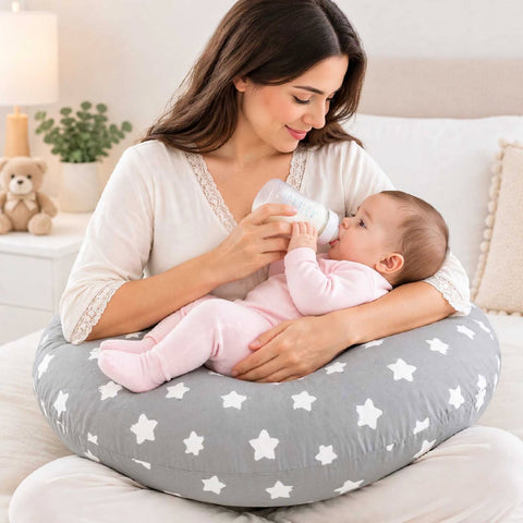 A woman sits on a white sofa using a gray nursing pillow with white stars to support a baby wearing a pink footed onesie while the baby drinks from a bottle.
