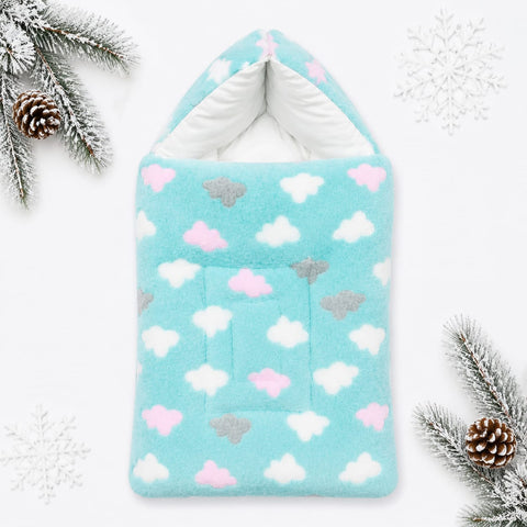 A soft fleece baby blanket or pouch with a hood, featuring a light blue background with white, pink, and gray cloud patterns. The product is styled in a flat-lay arrangement with white snowflakes and pine branches on a white background.