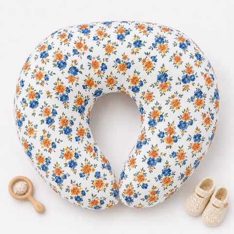 A U-shaped nursing pillow with a white background and a blue and orange floral pattern, styled with a wooden baby rattle and a pair of cream baby shoes on a white background.