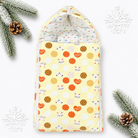 A baby swaddle blanket with a festive holiday print, featuring a pale yellow background with orange hearts, brown polka dots, white polka dots, and cute bunny faces. The swaddle is styled flat-lay with pine branches, pinecones, and white snowflake decorat