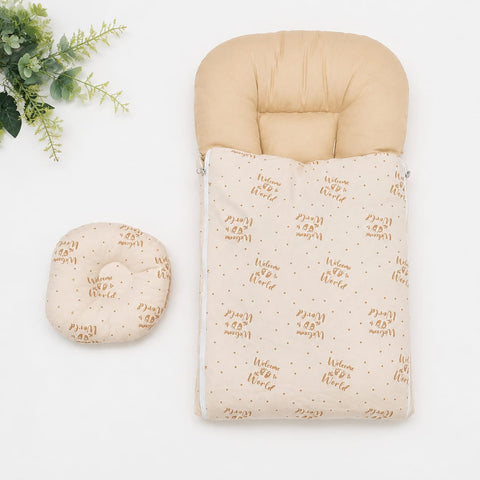 A baby sleep bag set with a beige padded hood and a cream fabric body printed with brown 'Welcome to the World' text and small brown dots, shown with a matching curved cushion on a white background with a small green plant in the corner.