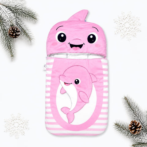 A pink baby blanket or sleeping bag with a shark hood featuring a smiling cartoon face and a pink dolphin illustration on the front, styled with white pine branches, snowflakes, and pinecones on a white background.